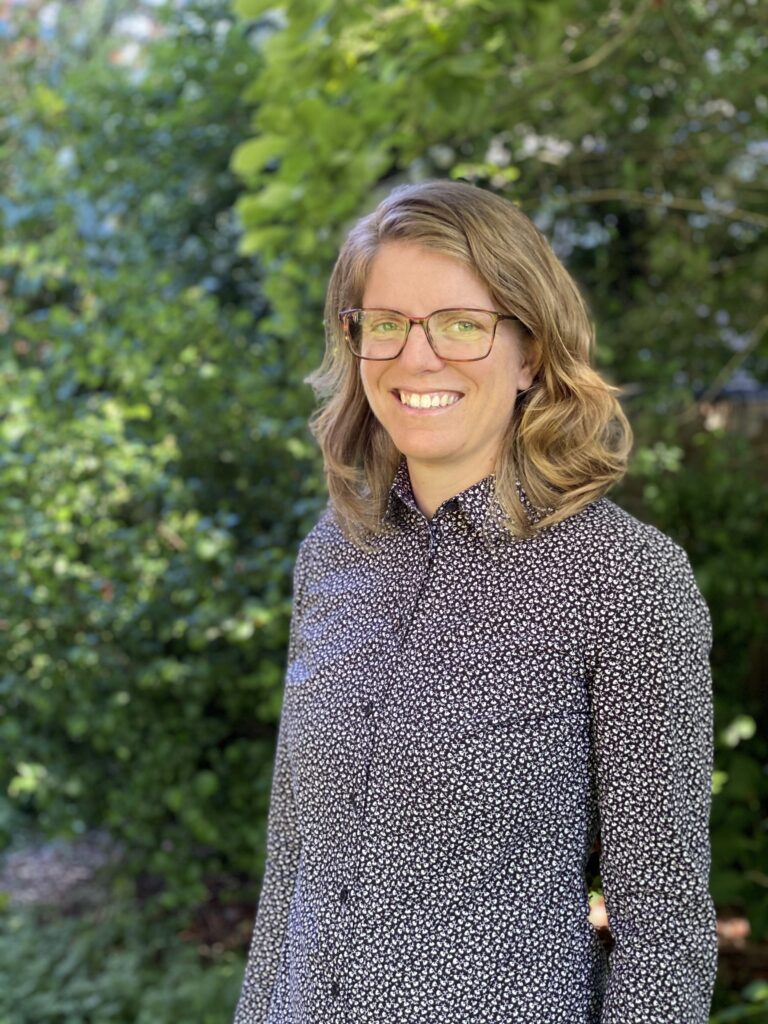 Cara Stoddard smiles at the camera in front of lush foliage background. They have blonde shoulder-length hair and glasses and are wearing a floral-pattern button-down shirt.